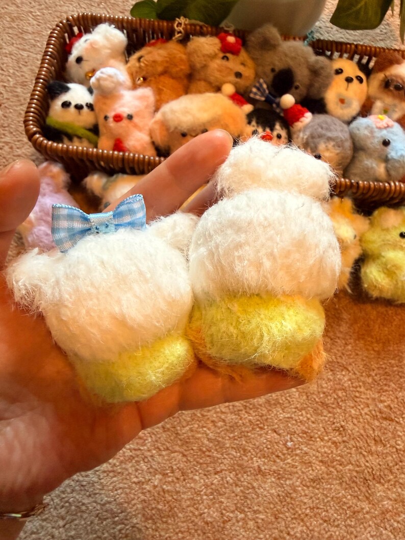 May include: A hand holding two small, fluffy, white and yellow plush toys with a blue and white checkered bow. A wicker basket filled with various small plush animals is in the background.