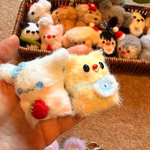 May include: A collection of handmade, fluffy animal figurines, including a panda, bear, and penguin, displayed in a woven basket. Two plush toys are held in a hand, one white with a strawberry, the other yellow with a blue bib.