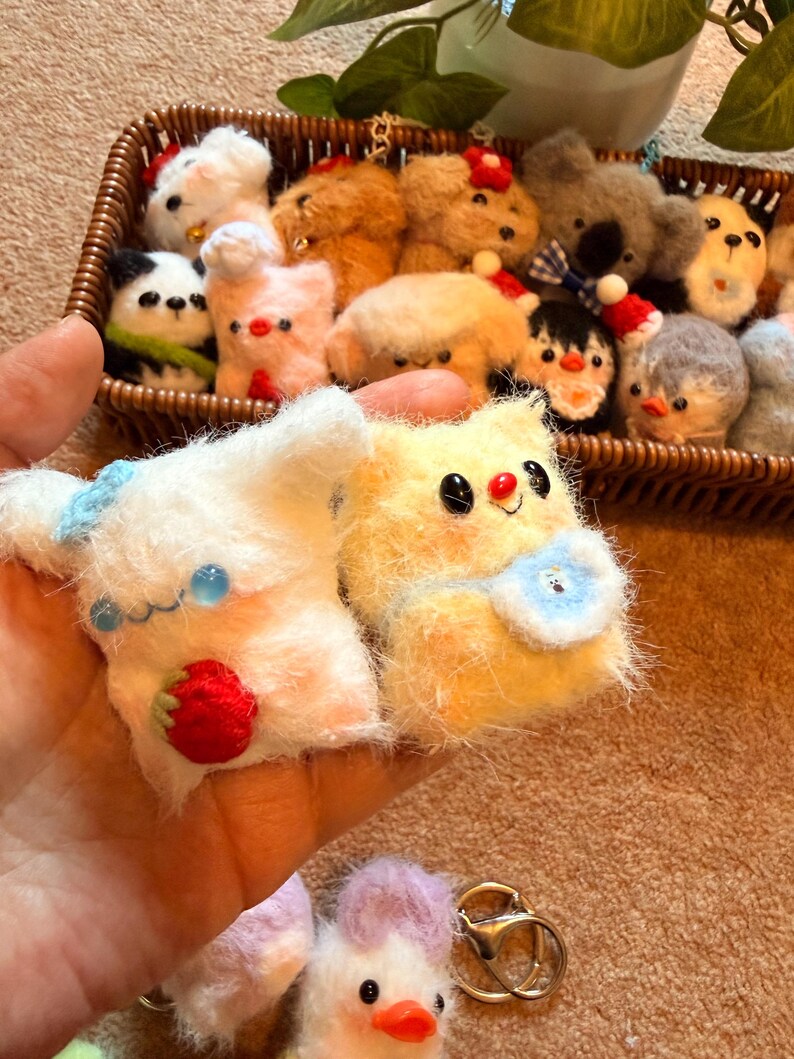 May include: A collection of handmade, fluffy animal figurines, including a panda, bear, and penguin, displayed in a woven basket. Two plush toys are held in a hand, one white with a strawberry, the other yellow with a blue bib.
