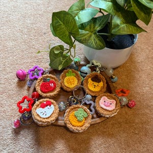 May include: A collection of handmade crochet keychains featuring various food designs, including a tomato, pineapple, and pig. The keychains are arranged on a wooden tray with a small potted plant in the background. Each keychain has a metal ring.