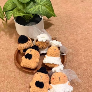May include: A collection of handmade crochet figures, some adorned with black hats and bowties, others with white bridal veils and dresses. The figures are arranged on a wooden plate, with a potted plant in the background.