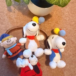 May include: Crocheted Snoopy and Woodstock figures in various poses. Snoopy wears a red collar, blue jacket, and holds balloons. Woodstock is in a red outfit. Other figures include a character in a blue hat and orange outfit. The figures are on a wooden tray.