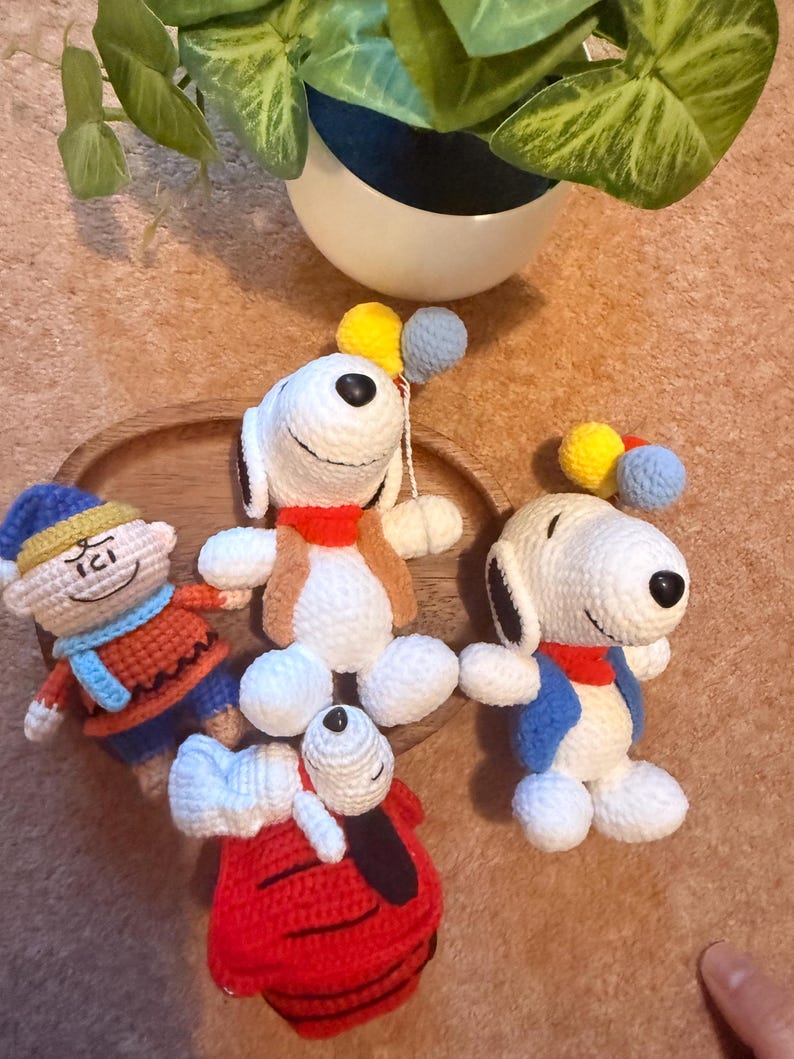 May include: Crocheted Snoopy and Woodstock figures in various poses. Snoopy wears a red collar, blue jacket, and holds balloons. Woodstock is in a red outfit. Other figures include a character in a blue hat and orange outfit. The figures are on a wooden tray.