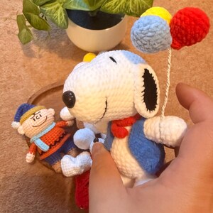 May include: Crocheted Snoopy doll with a blue vest, red scarf, and holding colorful pom-pom balloons. A smaller crocheted figure is also present. The toys are handmade and feature detailed stitching. The background includes a potted plant.