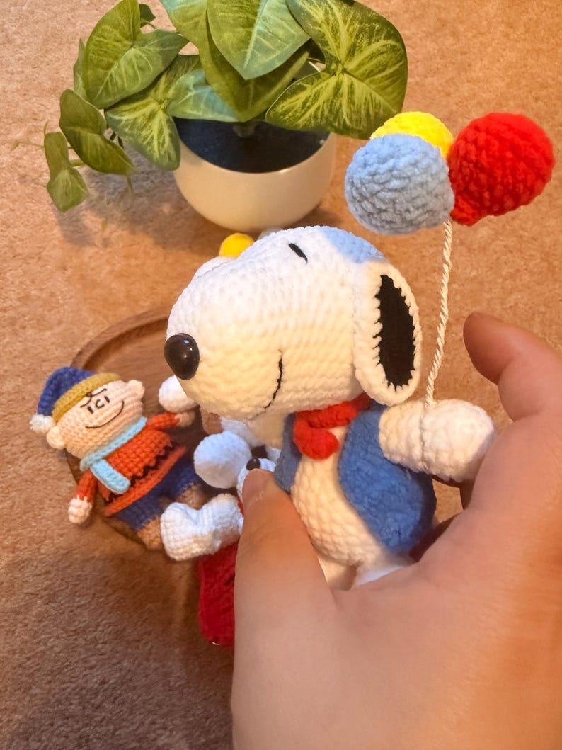 May include: Crocheted Snoopy doll with a blue vest, red scarf, and holding colorful pom-pom balloons. A smaller crocheted figure is also present. The toys are handmade and feature detailed stitching. The background includes a potted plant.