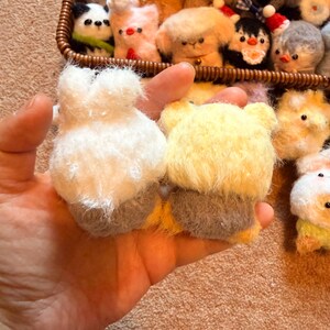 May include: A hand holds two small, handmade felt animal figures: a white bunny and a yellow bear. A basket in the background is filled with various felt animal ornaments, including a panda, penguin, and other creatures. The felt animals are soft and fluffy.