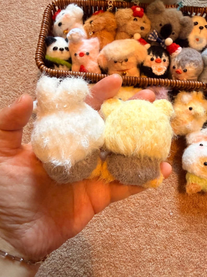 May include: A hand holds two small, handmade felt animal figures: a white bunny and a yellow bear. A basket in the background is filled with various felt animal ornaments, including a panda, penguin, and other creatures. The felt animals are soft and fluffy.