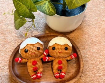 May include: Two crocheted gingerbread men with white icing hair, red and white striped arms and legs, and red button details. They are displayed on a wooden plate, with a potted plant in the background.