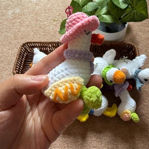Puede incluir: Un llavero de ganso hecho a mano con ganchillo, sostenido en una mano, con cuerpo blanco, sombrero rosa, bufanda morada y pico naranja. Otros llaveros de ganso similares est&aacute;n en una cesta. Los llaveros tienen detalles en amarillo, verde y azul.