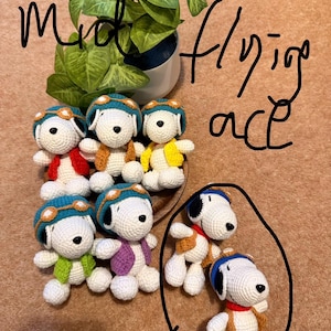May include: Handmade crochet Snoopy dolls, each wearing a pilot's cap and jacket in various colors. The dolls are white with black ears and a black spot. The image also includes a potted plant and handwritten text that says "mid flying ace stand et".