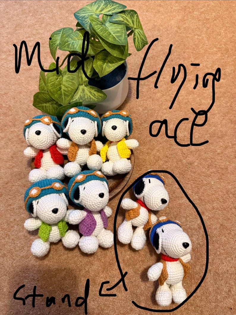 May include: Handmade crochet Snoopy dolls, each wearing a pilot's cap and jacket in various colors. The dolls are white with black ears and a black spot. The image also includes a potted plant and handwritten text that says "mid flying ace stand et".