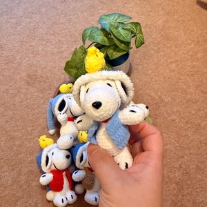 May include: Crocheted Snoopy dog dolls with blue and red vests and hats. A small yellow crocheted bird is perched on the head of each Snoopy doll. A small potted plant with green leaves is in the background.