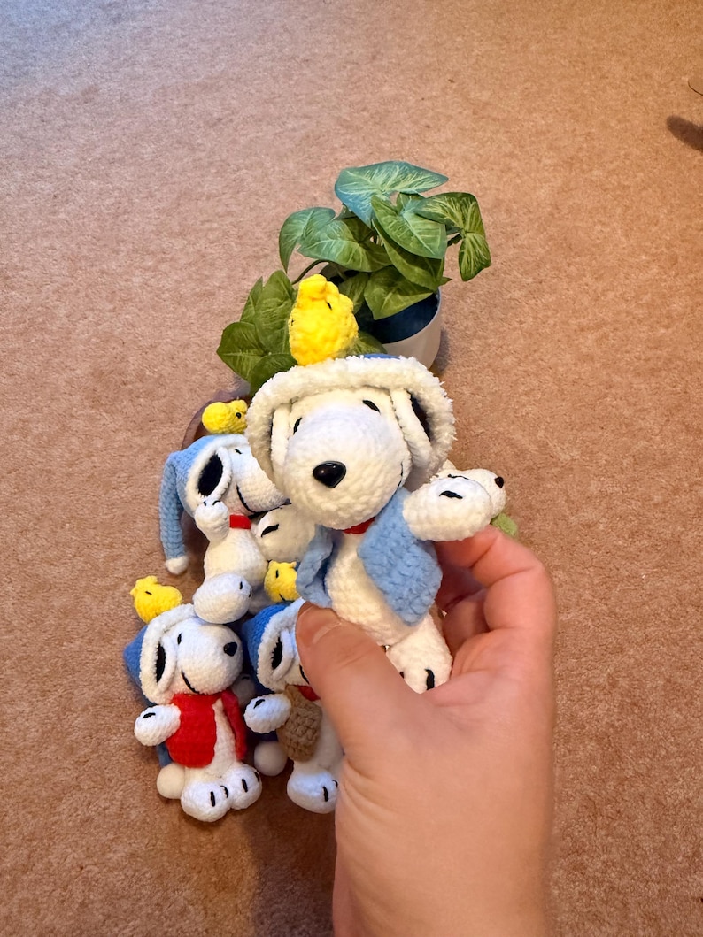 May include: Crocheted Snoopy dog dolls with blue and red vests and hats. A small yellow crocheted bird is perched on the head of each Snoopy doll. A small potted plant with green leaves is in the background.