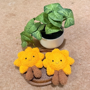 May include: Two plush yellow suns with brown legs and smiling faces sit on a wooden plate. A small white pot with green leafy plant sits behind the suns. The background is a light brown carpet.