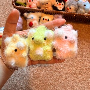 May include: A collection of handmade, fluffy animal keychains in a variety of colors. The keychains are primarily yellow, green, and pink, with small black eyes and white accents. A basket filled with similar plush animals is in the background.