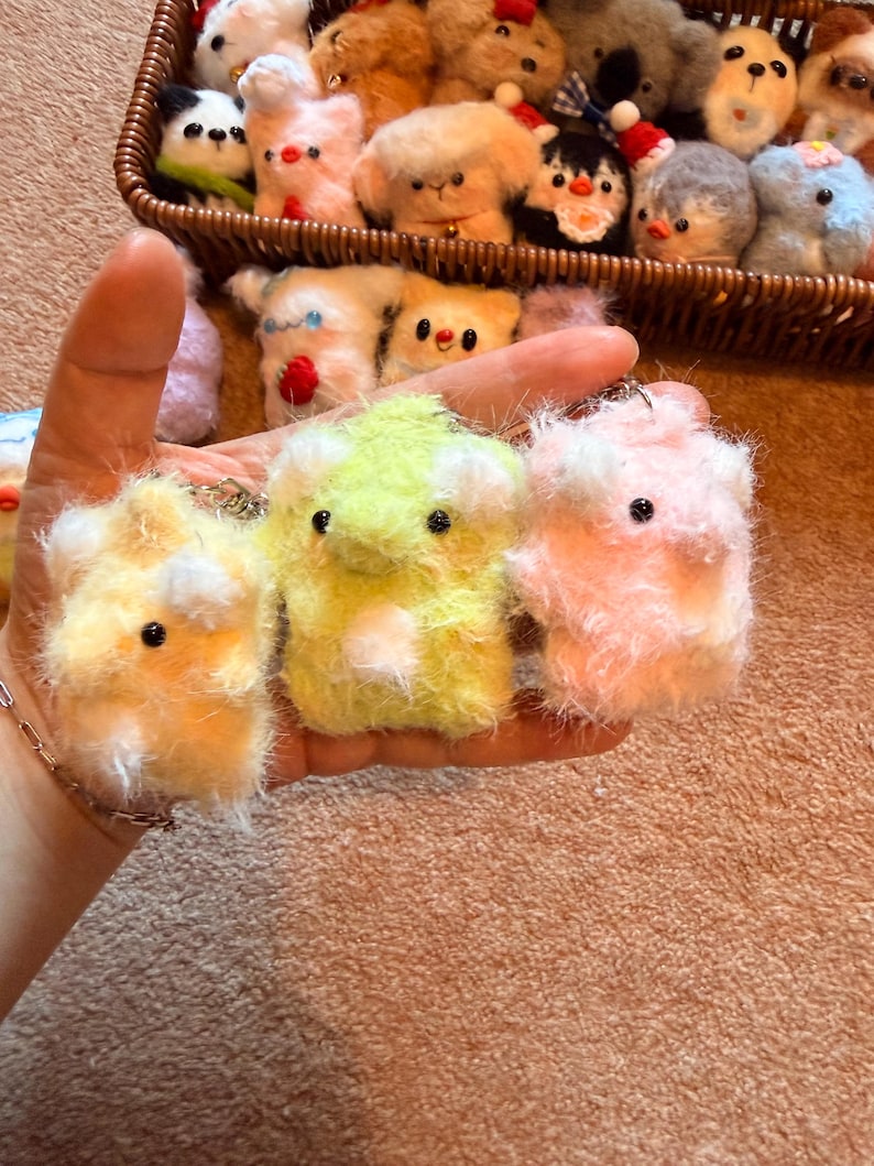 May include: A collection of handmade, fluffy animal keychains in a variety of colors. The keychains are primarily yellow, green, and pink, with small black eyes and white accents. A basket filled with similar plush animals is in the background.