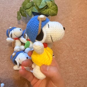 May include: Crocheted Snoopy dolls, featuring white bodies, black ears, and blue aviator hats with brown accents. Each doll has a red collar. One doll is held in a hand, while others are arranged nearby. A small potted plant is in the background.
