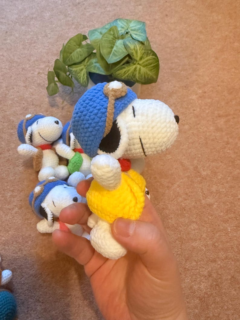 May include: Crocheted Snoopy dolls, featuring white bodies, black ears, and blue aviator hats with brown accents. Each doll has a red collar. One doll is held in a hand, while others are arranged nearby. A small potted plant is in the background.