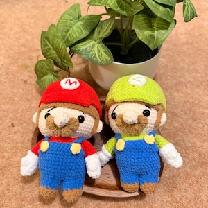 May include: Two handmade crochet figures of Mario and Luigi, iconic video game characters. Mario wears a red hat with a white 'M', blue overalls, and a brown mustache. Luigi has a green hat, blue overalls, and a brown mustache. They are on a wooden tray.
