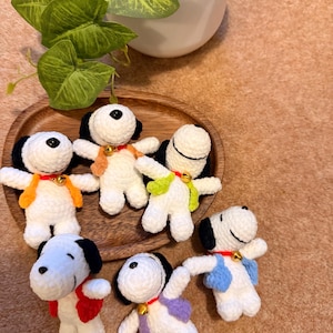May include: A collection of handmade crochet Snoopy figures. Each Snoopy has a white body, black ears, and a black spot on top of the head. They wear colorful vests and collars with small gold bells. The figures are arranged on a wooden tray.