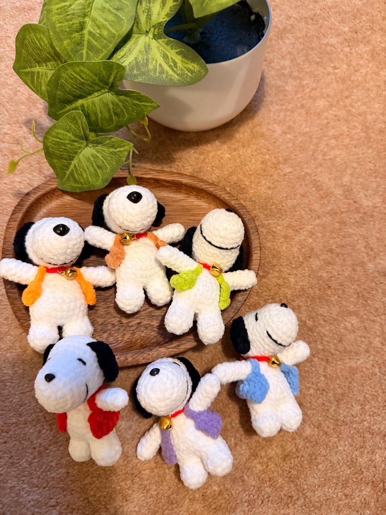 May include: A collection of handmade crochet Snoopy figures. Each Snoopy has a white body, black ears, and a black spot on top of the head. They wear colorful vests and collars with small gold bells. The figures are arranged on a wooden tray.
