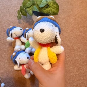 May include: Handmade crochet Snoopy dolls. Each doll is white with a blue and brown aviator hat, a red collar, and a yellow vest. The dolls are small and appear to be made of soft yarn. A green plant in a white and blue pot is in the background.