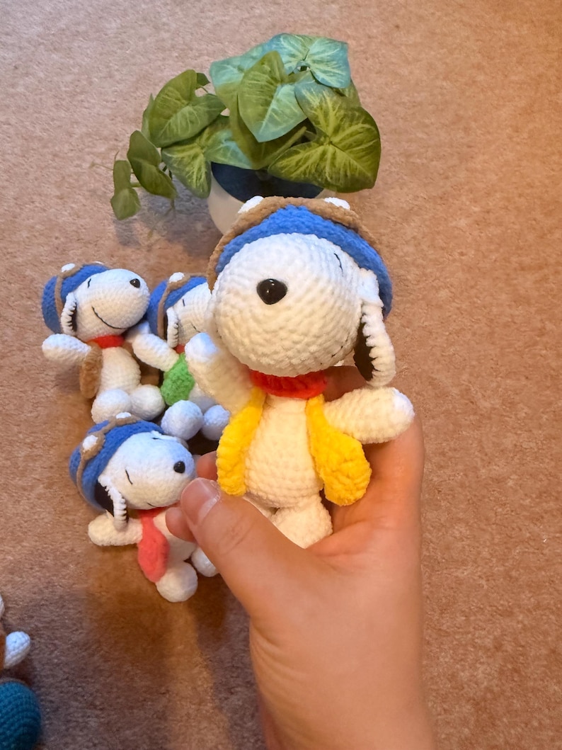 May include: Handmade crochet Snoopy dolls. Each doll is white with a blue and brown aviator hat, a red collar, and a yellow vest. The dolls are small and appear to be made of soft yarn. A green plant in a white and blue pot is in the background.