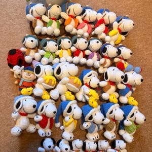May include: A collection of handmade Snoopy plush toys in various outfits. The crochet figures feature white bodies, black ears, and Snoopy's signature black nose. Each Snoopy wears a different colored hat and scarf, with some holding small yellow birds.