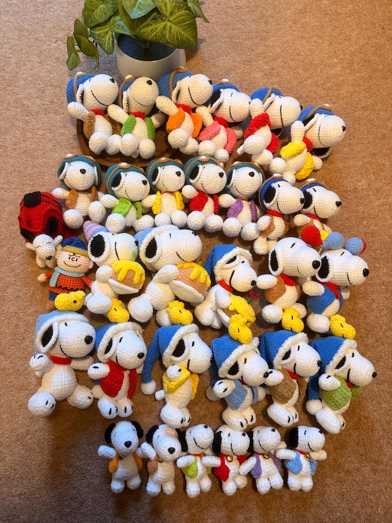 May include: A collection of handmade Snoopy plush toys in various outfits. The crochet figures feature white bodies, black ears, and Snoopy's signature black nose. Each Snoopy wears a different colored hat and scarf, with some holding small yellow birds.