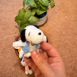 May include: A handmade Snoopy crochet doll, featuring a white body, black ears, and a black nose. The doll wears a blue scarf and a red collar with a golden bell. A small green plant in a white pot is in the background.