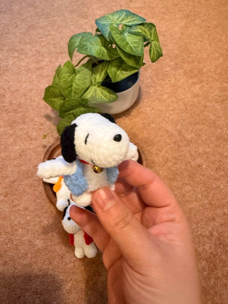 May include: A handmade Snoopy crochet doll, featuring a white body, black ears, and a black nose. The doll wears a blue scarf and a red collar with a golden bell. A small green plant in a white pot is in the background.