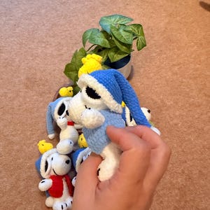 May include: Crocheted Snoopy dolls wearing blue nightcaps and outfits, with a red vest on one. Small yellow crocheted chicks are also present. A small potted plant is in the background. The dolls are white, blue, red, and yellow.