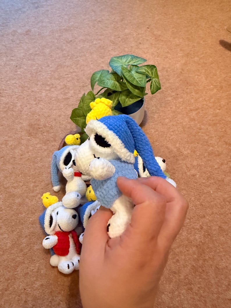 May include: Crocheted Snoopy dolls wearing blue nightcaps and outfits, with a red vest on one. Small yellow crocheted chicks are also present. A small potted plant is in the background. The dolls are white, blue, red, and yellow.