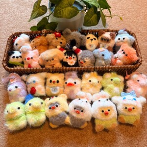 May include: A collection of handmade felt animal keychains in a woven basket. The keychains include various animals such as dogs, birds, and koalas, in a variety of colors including white, brown, yellow, and blue. A potted plant is in the background.