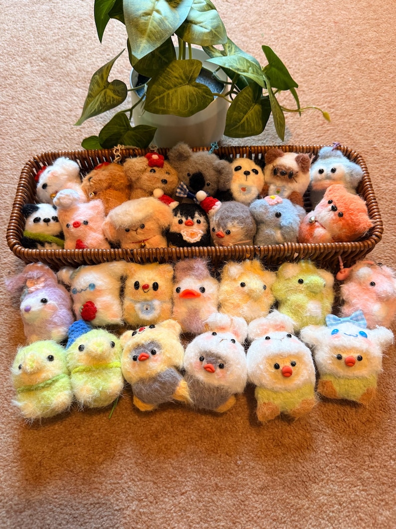 May include: A collection of handmade felt animal keychains in a woven basket. The keychains include various animals such as dogs, birds, and koalas, in a variety of colors including white, brown, yellow, and blue. A potted plant is in the background.