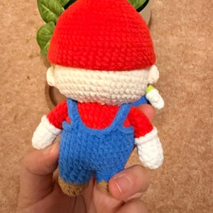 May include: A handmade crochet doll of a popular video game character. The doll features a red hat, red shirt, blue overalls, and white gloves. The doll is held in a person's hand, with a plant in the background.