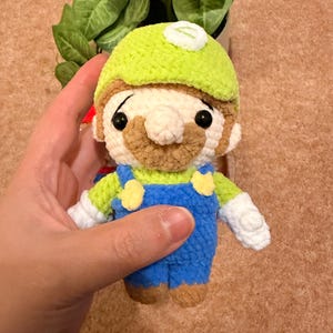 May include: A handmade crochet doll of a character wearing a green hat, blue overalls, and a brown mustache. The doll has black eyes and white gloves. The character is a popular video game character.