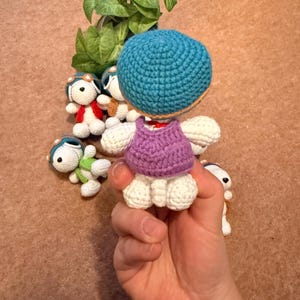 May include: A collection of handmade crochet dolls. The main doll is held in a hand and features a blue head, purple top, and white body. Other dolls in the background have aviator hats and various colored outfits. The dolls are on a brown surface.