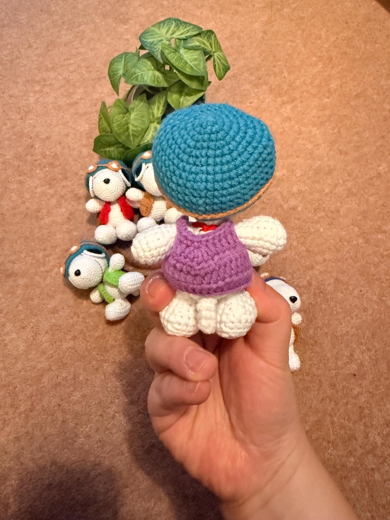May include: A collection of handmade crochet dolls. The main doll is held in a hand and features a blue head, purple top, and white body. Other dolls in the background have aviator hats and various colored outfits. The dolls are on a brown surface.