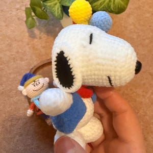 May include: Crocheted Snoopy doll with a blue vest, red collar, and black ears. The doll has yellow and blue pom-poms on top of its head. A small crocheted figure of a character in a blue hat is also visible.