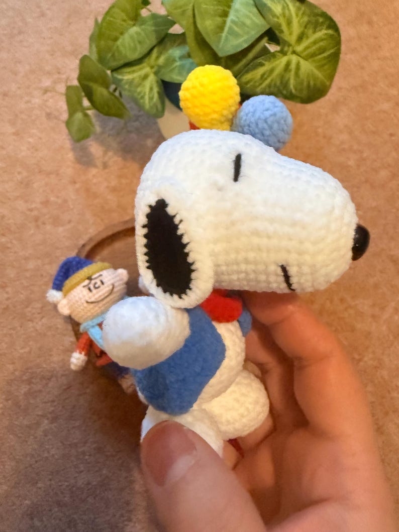 May include: Crocheted Snoopy doll with a blue vest, red collar, and black ears. The doll has yellow and blue pom-poms on top of its head. A small crocheted figure of a character in a blue hat is also visible.