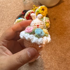 Pu&ograve; includere: Un portachiavi a forma di coniglietto all'uncinetto fatto a mano con un design a cupcake. Il coniglietto &egrave; bianco con accenti floreali rosa e blu. La base della cupcake &egrave; beige con una parte superiore a balze bianca. Altri oggetti all'uncinetto sono sullo sfondo.