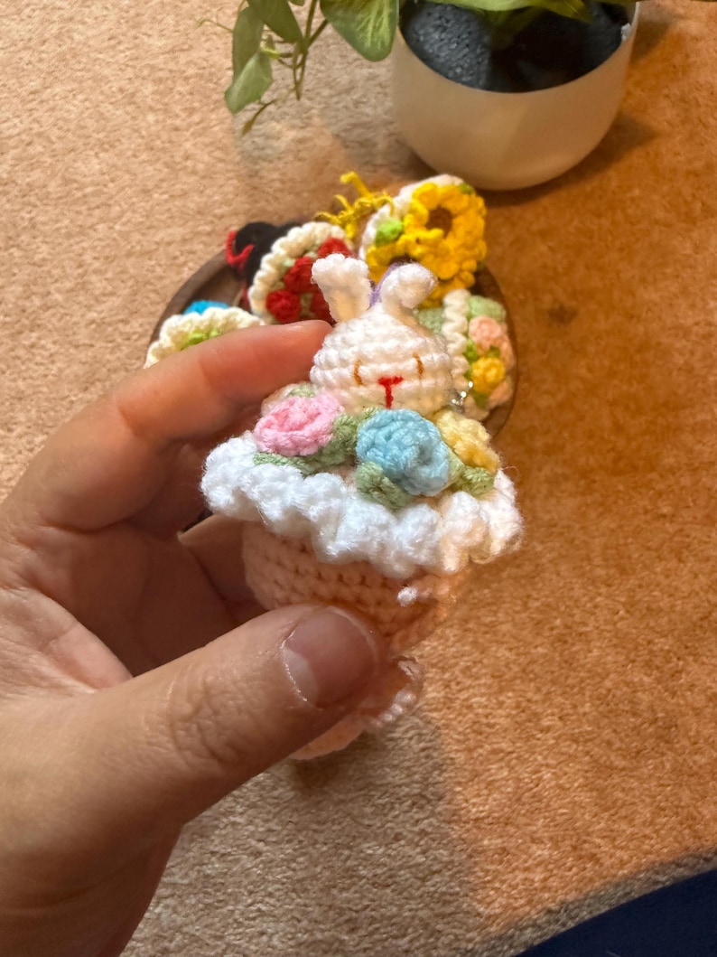 Pu&ograve; includere: Un portachiavi a forma di coniglietto all'uncinetto fatto a mano con un design a cupcake. Il coniglietto &egrave; bianco con accenti floreali rosa e blu. La base della cupcake &egrave; beige con una parte superiore a balze bianca. Altri oggetti all'uncinetto sono sullo sfondo.