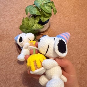 May include: Crocheted Snoopy figures, one holding a yellow cake with orange frosting. Snoopy wears a blue and pink party hat. A small potted plant with green leaves is in the background.