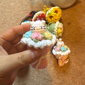Pu&ograve; includere: Portachiavi a forma di cupcake all'uncinetto con faccina di coniglietto, decorato con fiori colorati e una parte superiore bianca a balze. Il portachiavi &egrave; rosa e bianco, con un piccolo portachiavi cupcake abbinato nelle vicinanze. Lo sfondo &egrave; marrone neutro.
