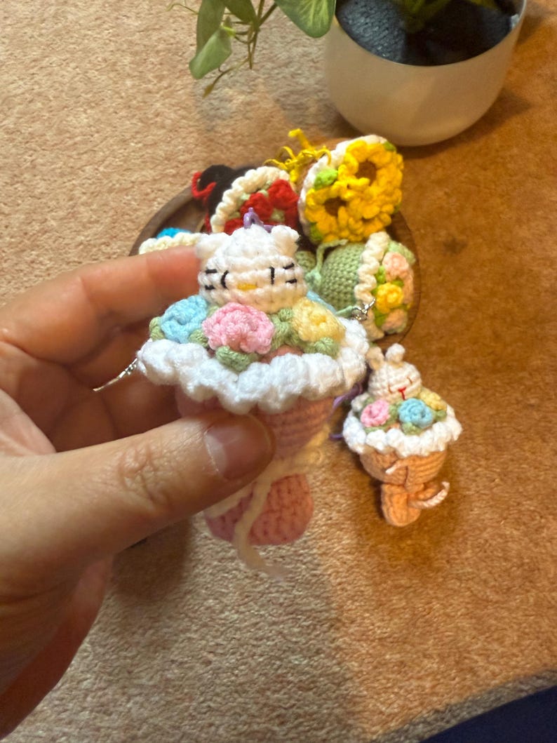 Pu&ograve; includere: Portachiavi a forma di cupcake all'uncinetto con faccina di coniglietto, decorato con fiori colorati e una parte superiore bianca a balze. Il portachiavi &egrave; rosa e bianco, con un piccolo portachiavi cupcake abbinato nelle vicinanze. Lo sfondo &egrave; marrone neutro.