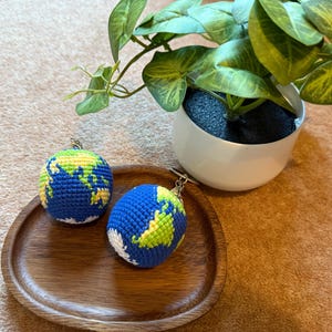 Puede incluir: Dos llaveros de globo de ganchillo azul y verde descansan sobre una bandeja de madera. Los llaveros presentan continentes amarillos y verdes, y los océanos son azules. Una planta en maceta blanca con hojas verdes está en el fondo.