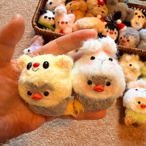 May include: Hand holding two small, handmade felt animal ornaments. One is yellow with a red nose and black eyes, the other is white with pink cheeks and a red beak. A basket in the background contains more felt animal ornaments.