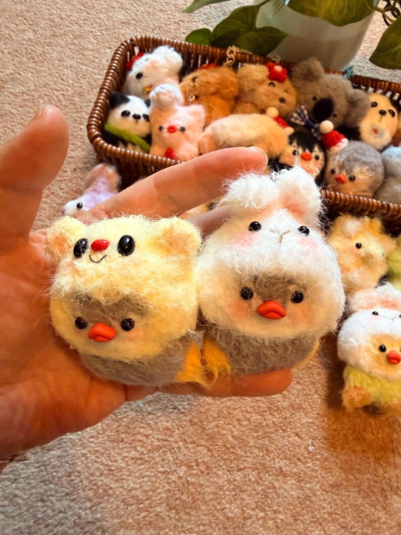 May include: Hand holding two small, handmade felt animal ornaments. One is yellow with a red nose and black eyes, the other is white with pink cheeks and a red beak. A basket in the background contains more felt animal ornaments.