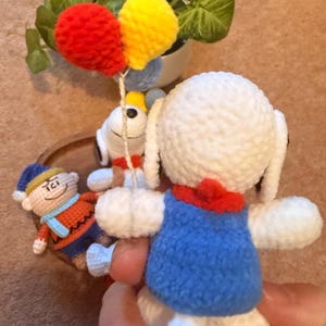 May include: Handmade crochet Snoopy doll wearing a blue vest and red collar. The doll is white with black eyes and ears. It is holding a string with red and yellow crochet balloons. A small crochet figure of Charlie Brown is in the background.